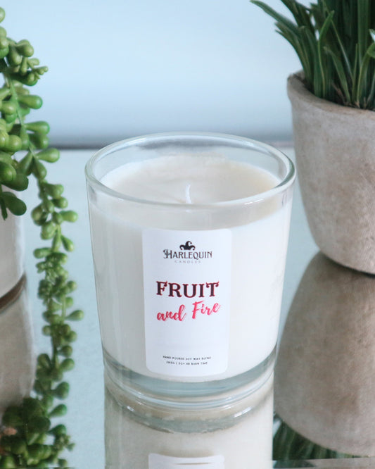 A Harlequin Candles "Fruit and Fire" hand-poured soy wax candle in a clear glass jar, displayed on a reflective surface next to potted green plants. Premium Australian made scented candle.