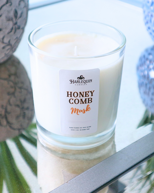 A Harlequin Candles "Honeycomb Musk" hand-poured soy wax candle in a clear glass jar, displayed on a reflective surface with decorative elements in the background. Premium Australian made scented candle.