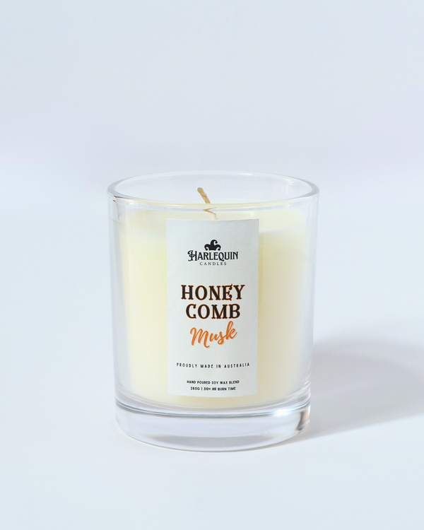 A Harlequin Candles "Honeycomb Musk" hand-poured soy wax candle in a clear glass jar, displayed on a reflective surface with decorative elements in the background. Premium Australian made scented candle.