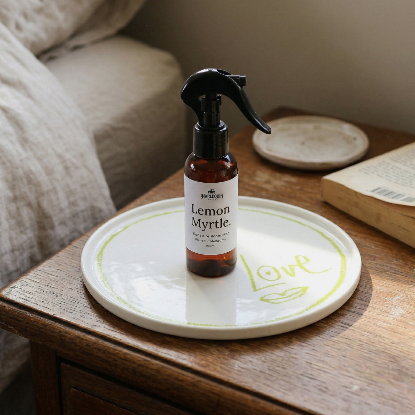 A bottle of Harlequin Candles Lemon Myrtle Signature Room Mist sitting on a decorative white tray on a wooden bedside table. This natural room spray is a beautiful home fragrance addition, perfect for bringing personality to your room.