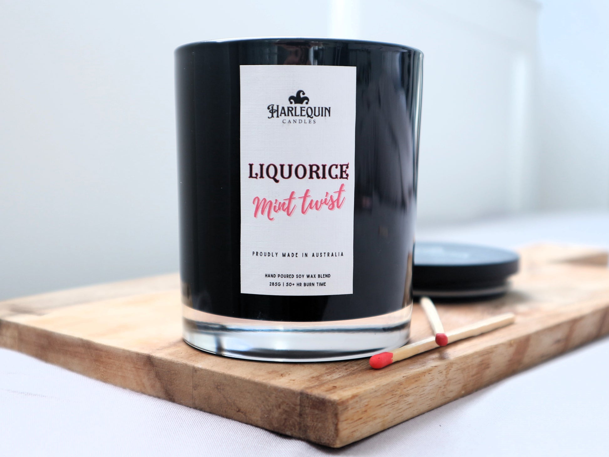 A Harlequin Candles "Liquorice Mint Twist" hand-poured soy blend candle in a sleek black glass jar, displayed on a wooden board with matches and its lid. Proudly Australian made.