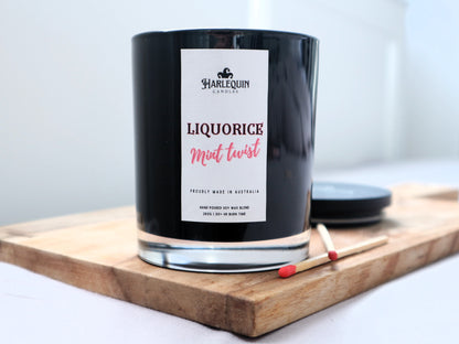 A Harlequin Candles "Liquorice Mint Twist" hand-poured soy blend candle in a sleek black glass jar, displayed on a wooden board with matches and its lid. Proudly Australian made.