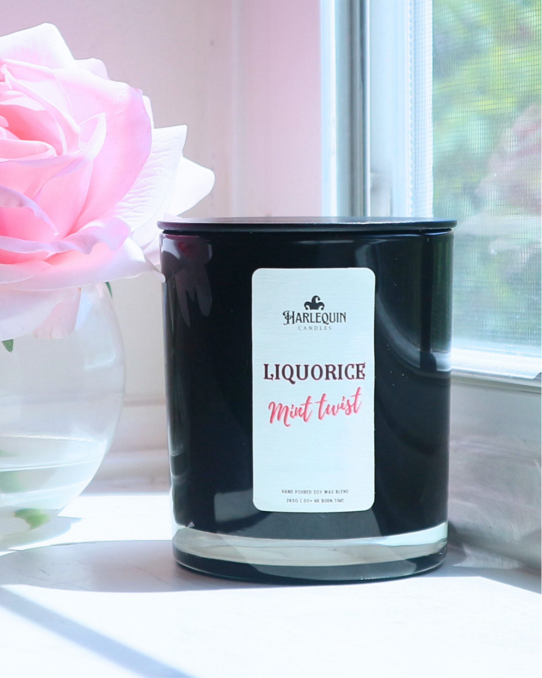 A Harlequin Candles "Liquorice Mint Twist" hand-poured soy wax candle in a black glass jar, styled beside a clear glass vase with vibrant pink roses. Premium Australian made scented candle.