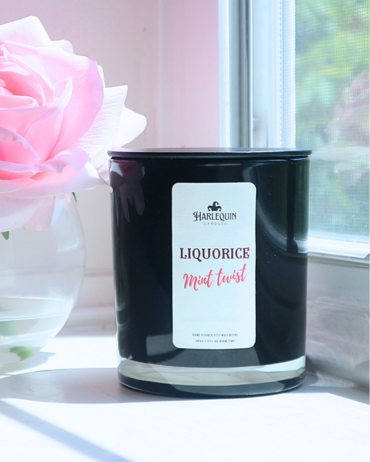 A Harlequin Candles "Liquorice Mint Twist" hand-poured soy wax candle in a black glass jar, styled beside a clear glass vase with vibrant pink roses. Premium Australian made scented candle.