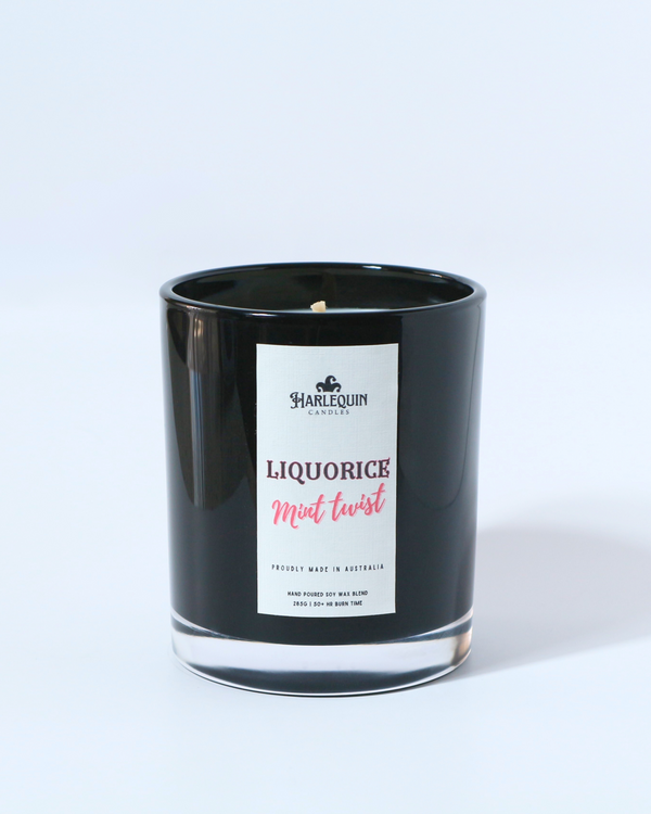 A Harlequin Candles "Liquorice Mint Twist" hand-poured soy wax candle in a black glass jar, styled beside a clear glass vase with vibrant pink roses. Premium Australian made scented candle.