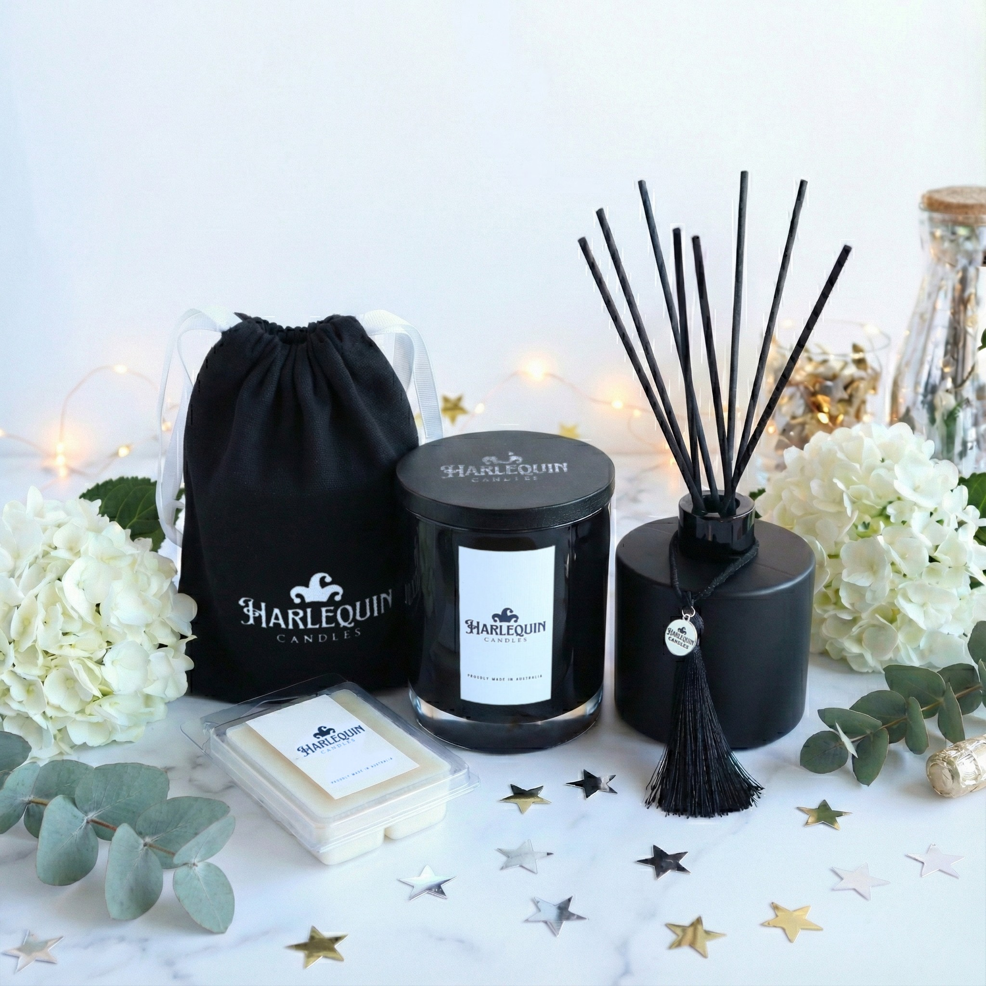Handmade Australian scented candle and reed diffuser gift set by Harlequin Candles. Includes a black jar candle, wax melts, and a branded drawstring gift bag, styled with fairy lights and white flowers for home decor inspiration