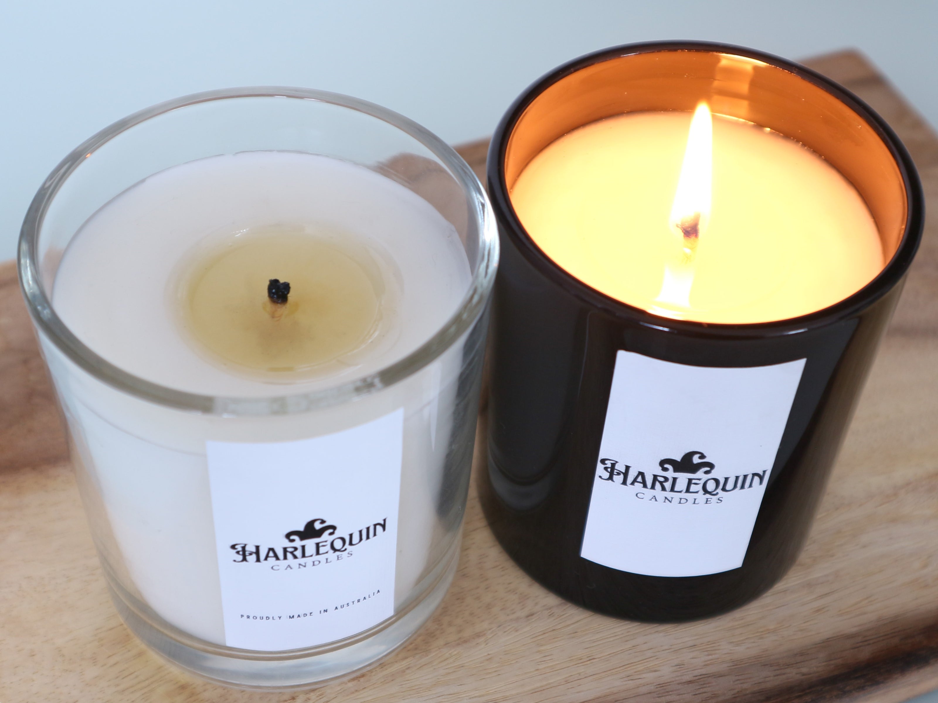Two Harlequin luxury scented candles on wood: one clear glass with white wax, the other black glass with a lit amber flame. Ideal home fragrance.