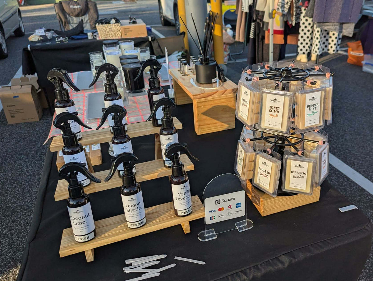 A close-up of the Harlequin Candles market stall display at Bentleigh Sunday Market, showcasing handmade soy wax melts, room mists, and reed diffusers made in Melbourne.