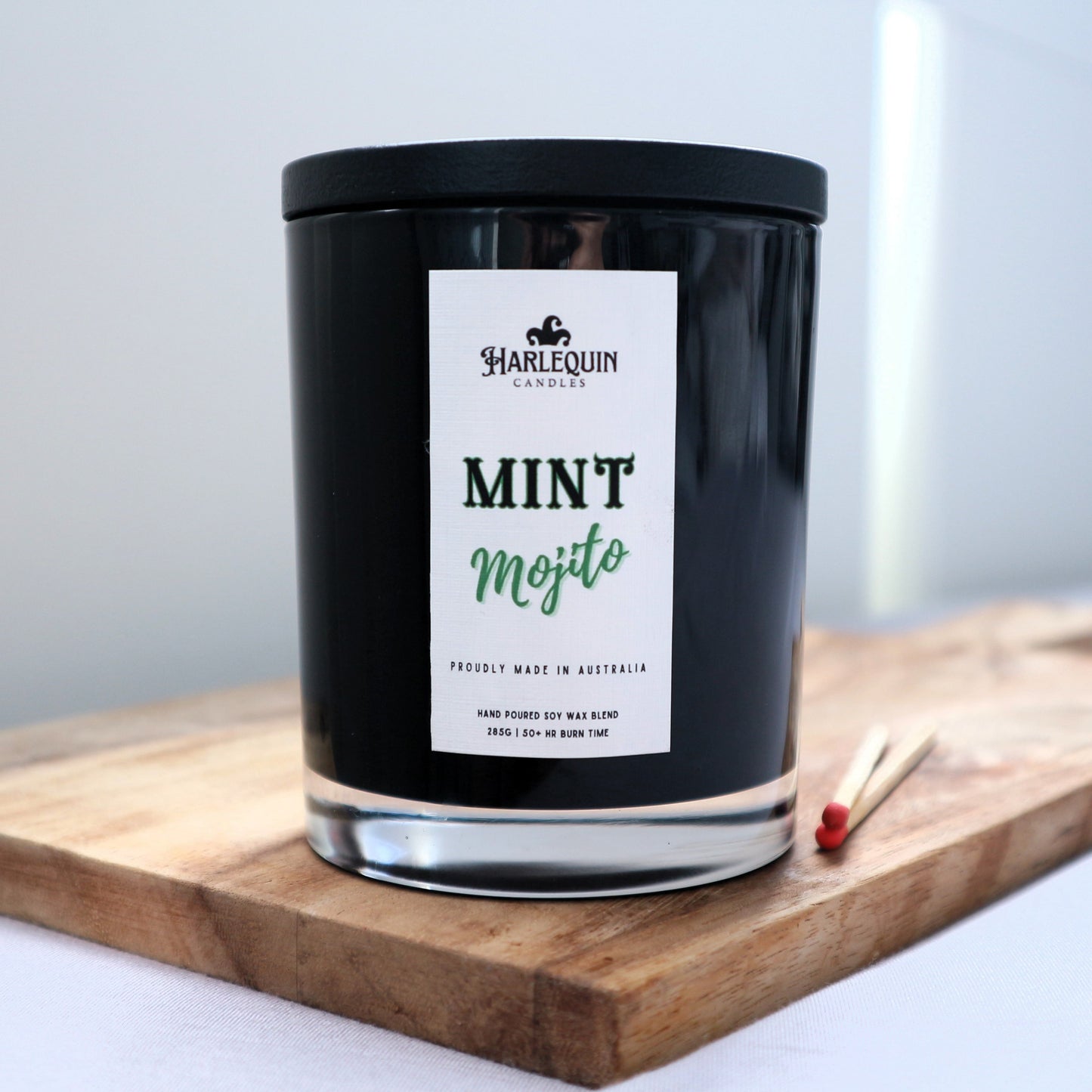 A Harlequin Candles "Mint Mojito" hand-poured soy blend candle in a sleek black glass jar with lid, displayed on a wooden board with matches. Proudly Australian made.