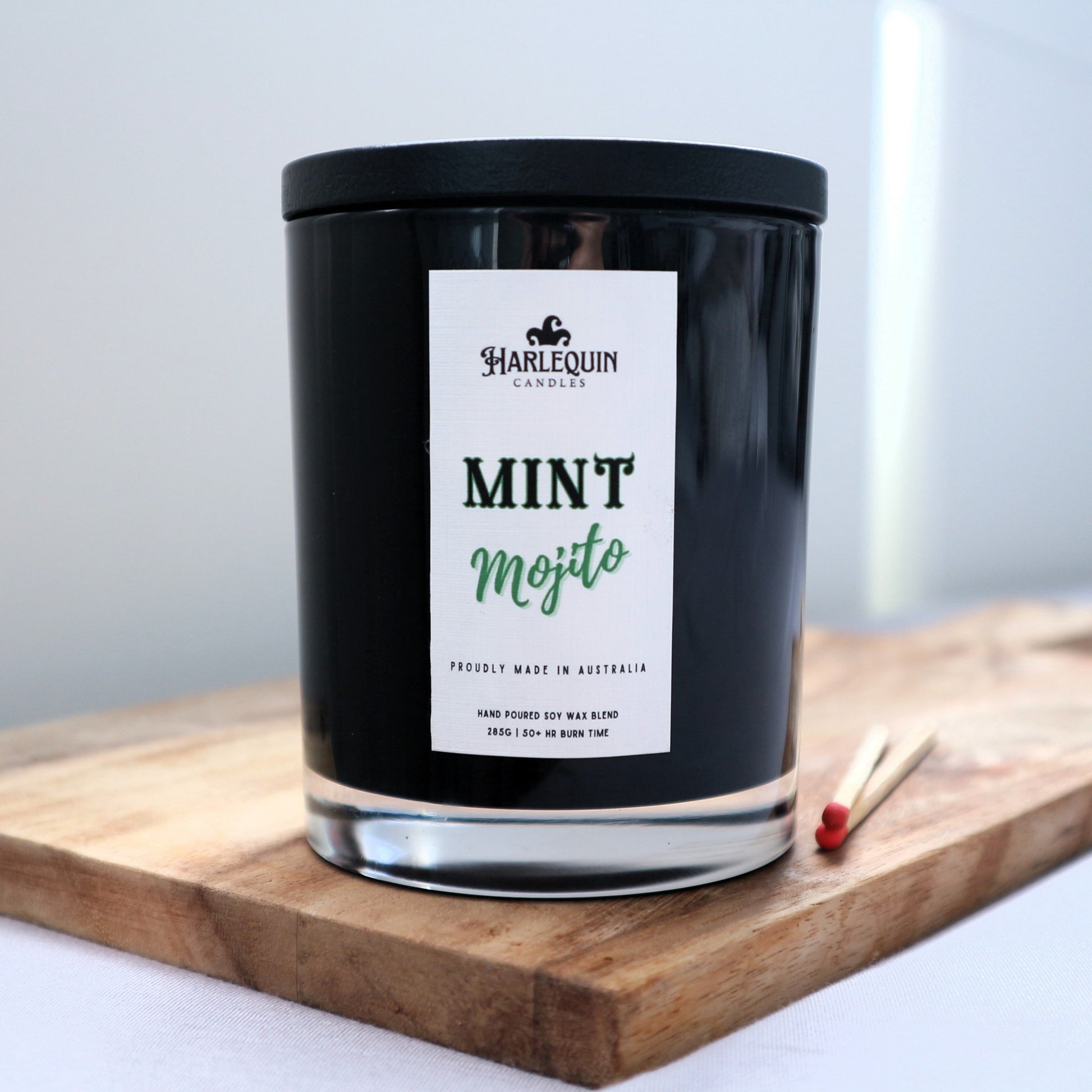 A Harlequin Candles "Mint Mojito" hand-poured soy blend candle in a sleek black glass jar with lid, displayed on a wooden board with matches. Proudly Australian made.