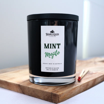 A Harlequin Candles "Mint Mojito" hand-poured soy blend candle in a sleek black glass jar with lid, displayed on a wooden board with matches. Proudly Australian made.