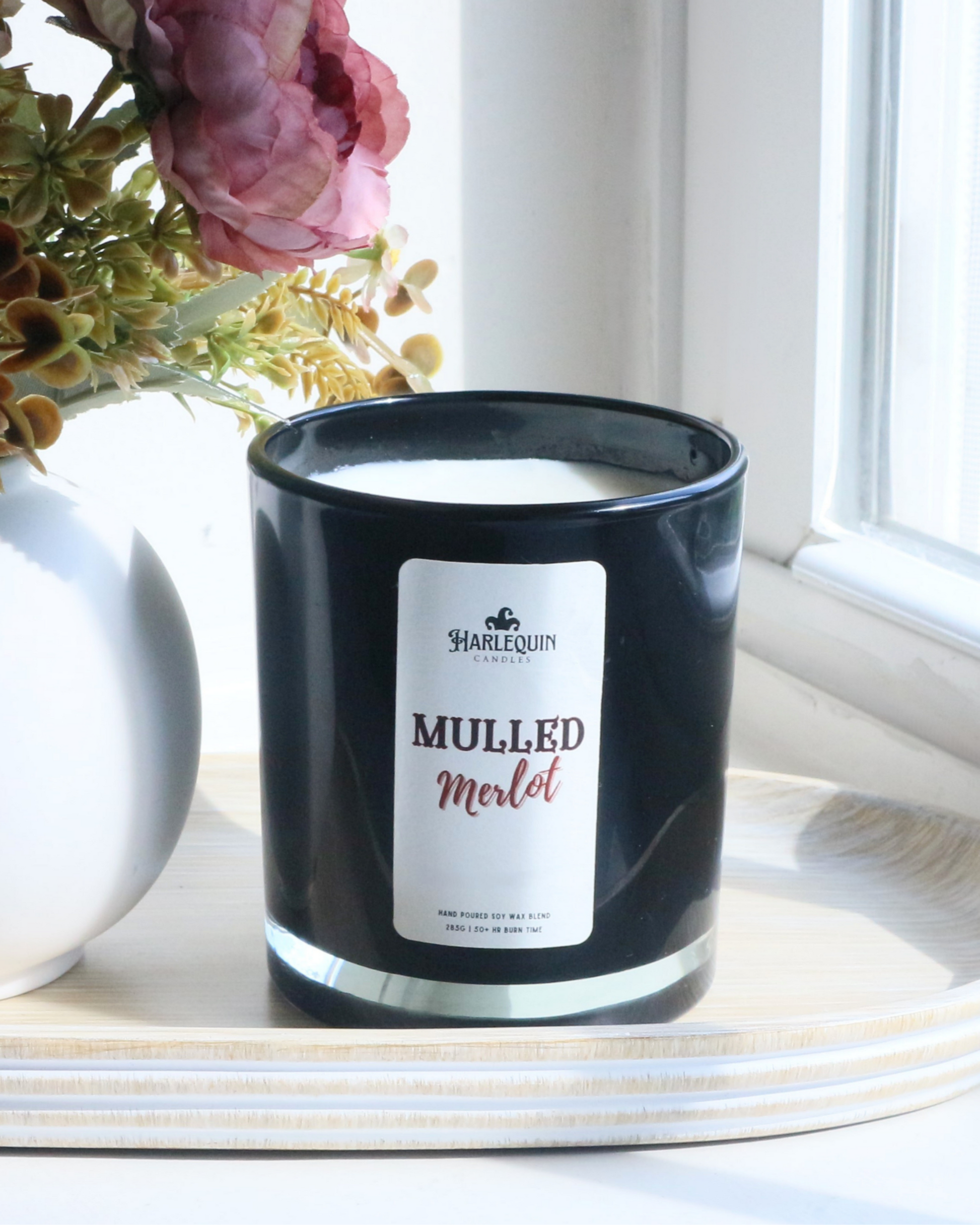 A Harlequin Candles "Mulled Merlot" hand-poured soy wax candle in a black glass jar, styled on a wooden tray beside a white vase with decorative flowers. Premium Australian made scented candle.