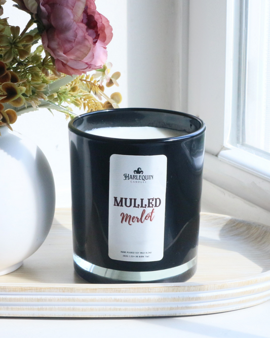 A Harlequin Candles "Mulled Merlot" hand-poured soy wax candle in a black glass jar, styled on a wooden tray beside a white vase with decorative flowers. Premium Australian made scented candle.