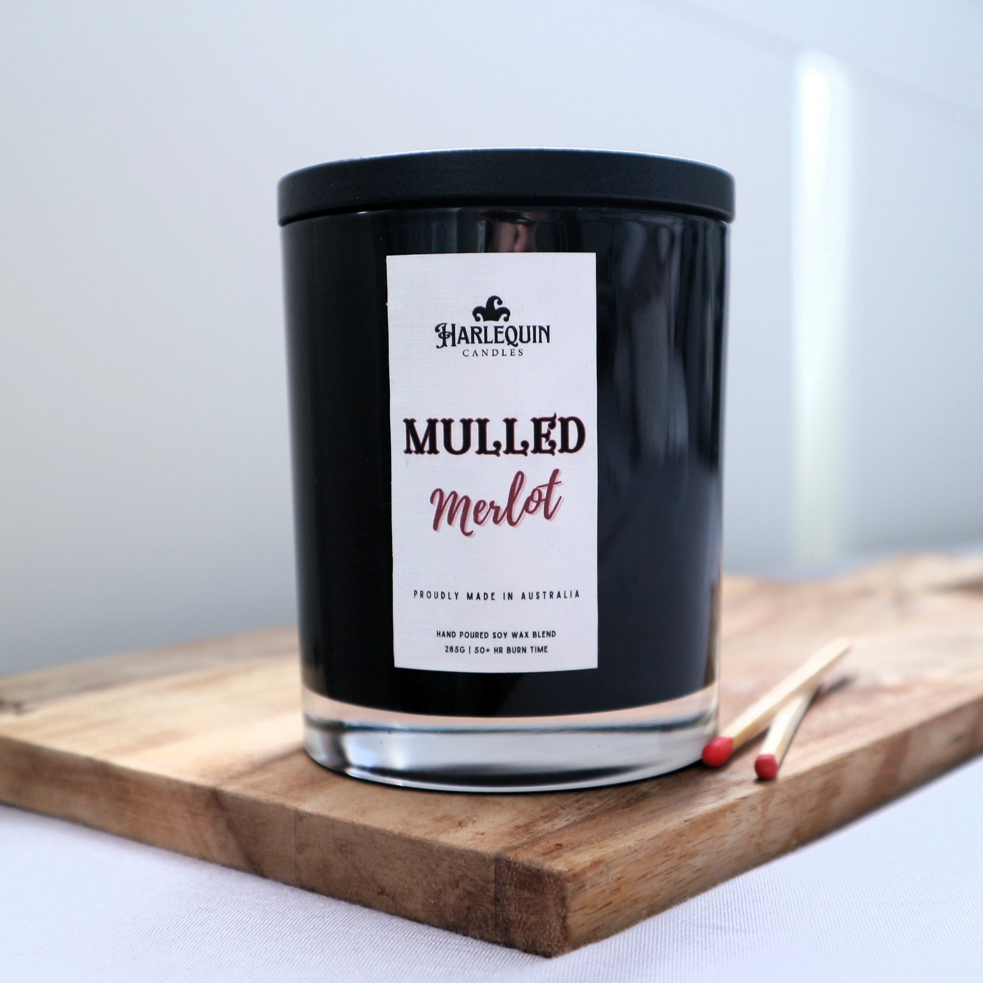 A Harlequin Candles "Mulled Wine" hand-poured soy blend candle in a sleek black glass jar with lid, displayed on a wooden board with matches. Proudly Australian made.