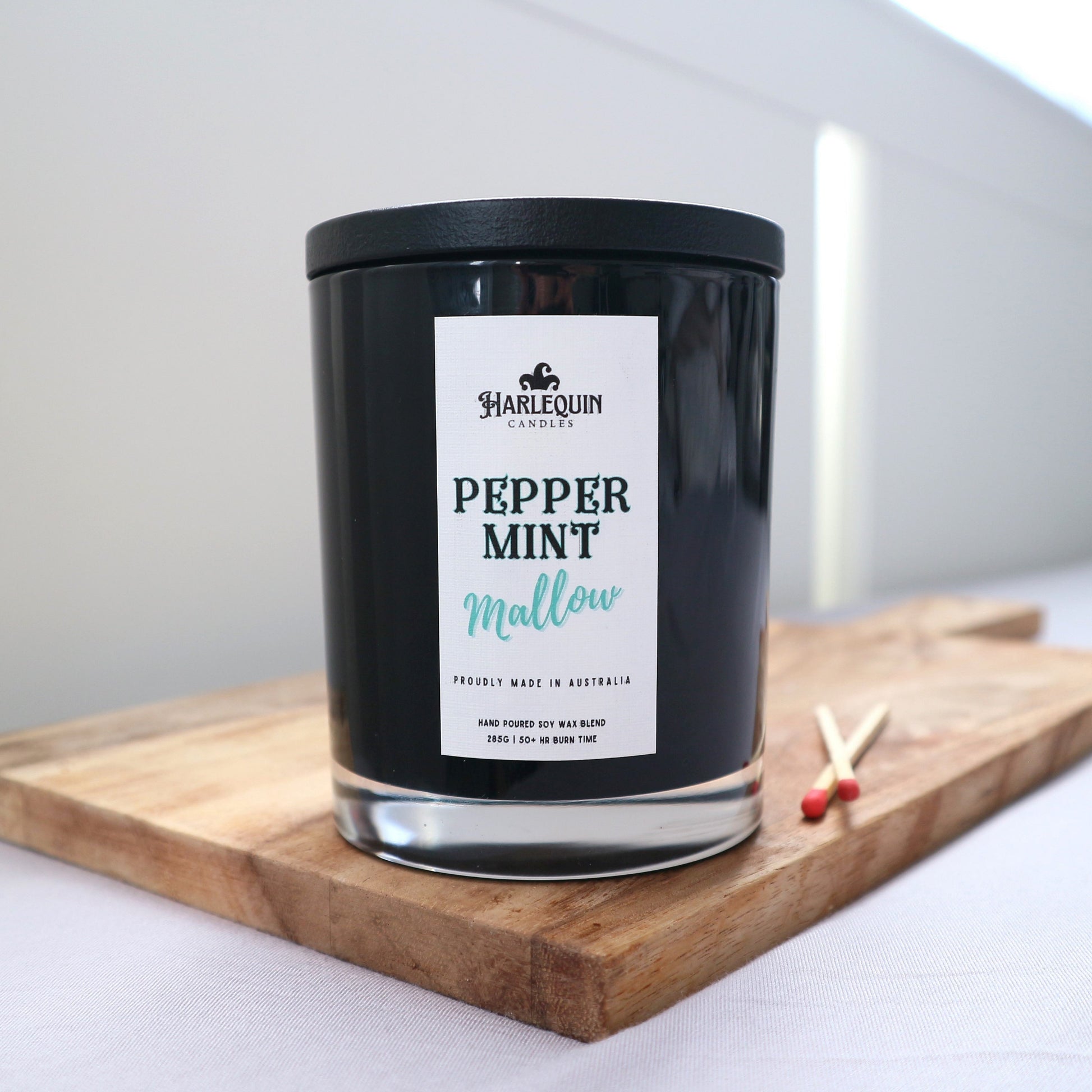 A Harlequin Candles "Peppermint Mallow" hand-poured soy blend candle in a sleek black glass jar with lid, displayed on a wooden board with matches. Proudly Australian made.