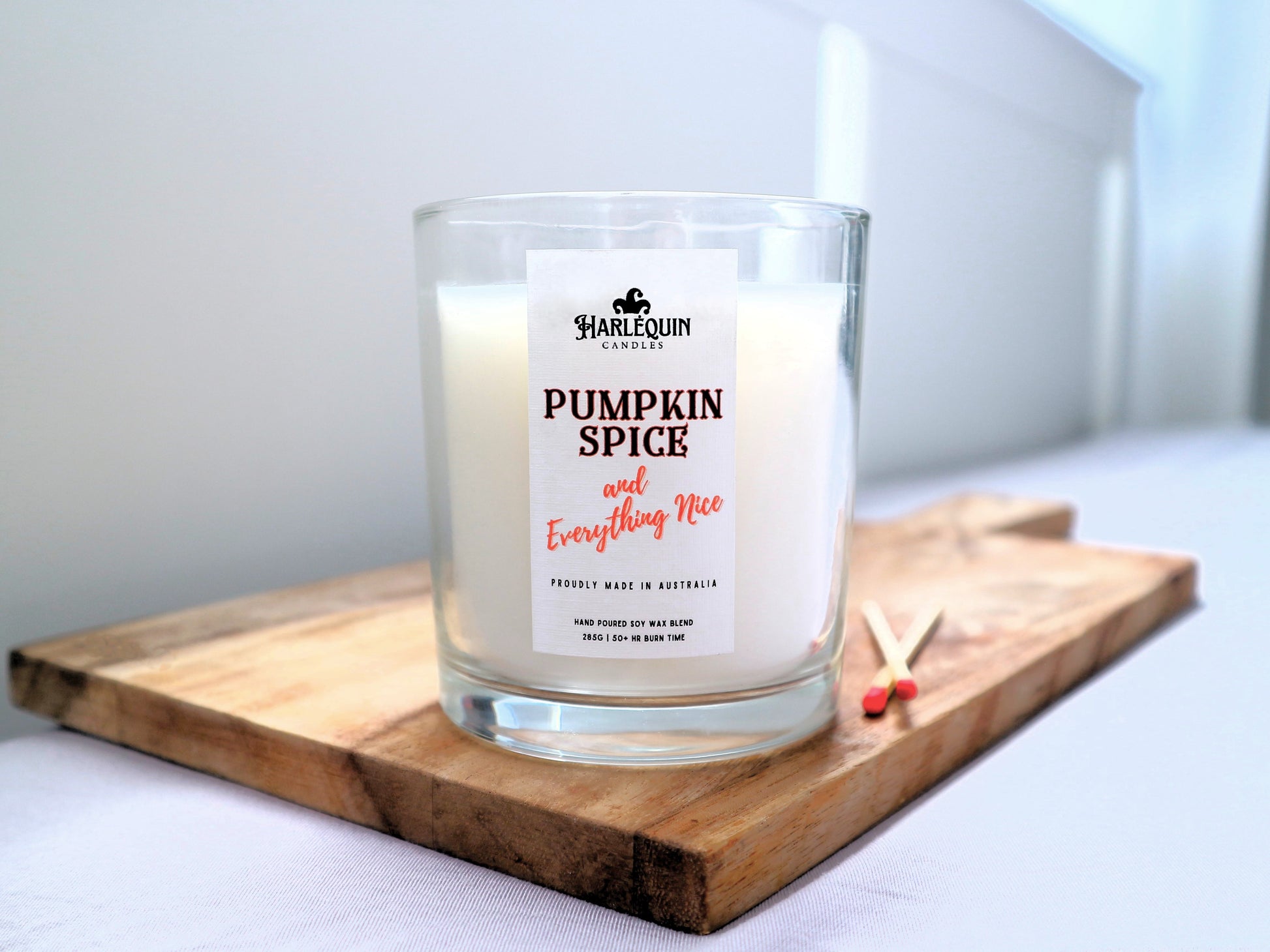 A Harlequin Candles "Pumpkin Spice and Everything Nice" hand-poured soy blend candle in a clear glass jar, displayed on a wooden board with matches. Proudly Australian made.