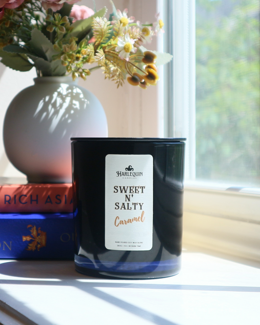 A Harlequin Candles "Sweet N' Salty Caramel" hand-poured soy wax candle in a black glass jar, styled beside a white vase with flowers and stacked books. Premium Australian made scented candle.