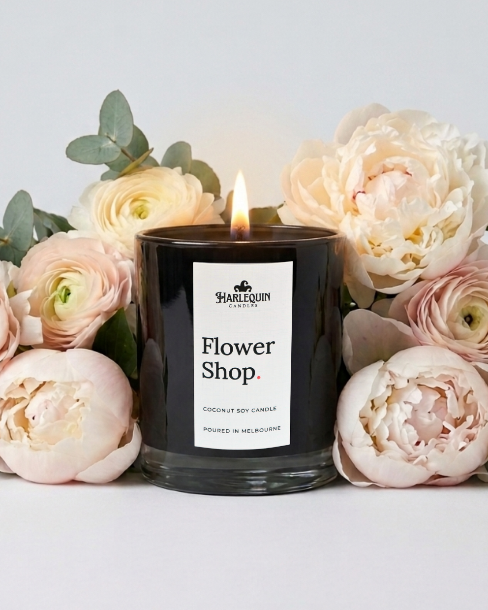 A beautifully lit, black Harlequin Candles "Flower Shop" Coconut Soy Candle, handpoured in Melbourne. Surrounded by soft cream peonies and roses, this artisan-made candle is an elegant gift idea for Mother’s Day 2026.