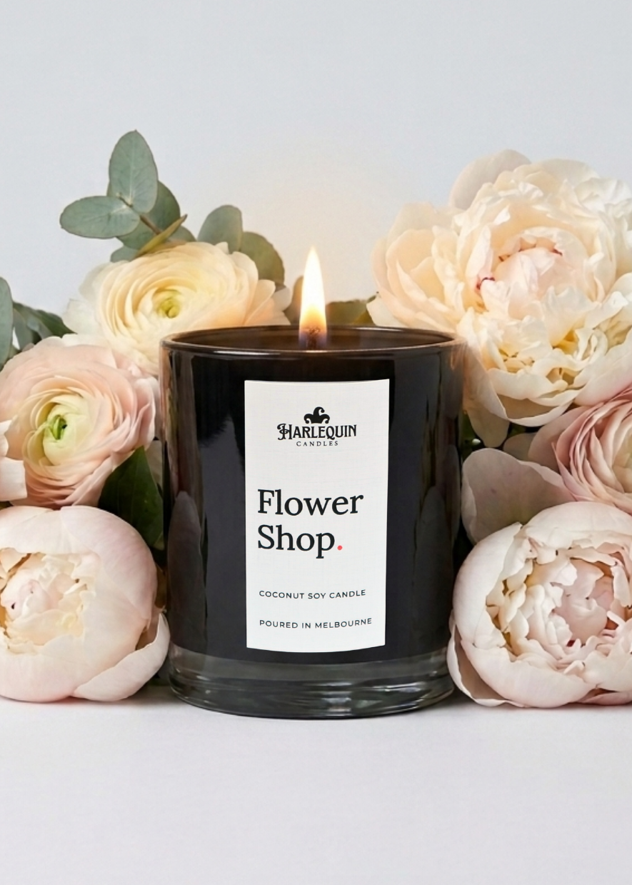 A beautifully lit, black Harlequin Candles "Flower Shop" Coconut Soy Candle, handpoured in Melbourne. Surrounded by soft cream peonies and roses, this artisan-made candle is an elegant gift idea for Mother’s Day 2026.