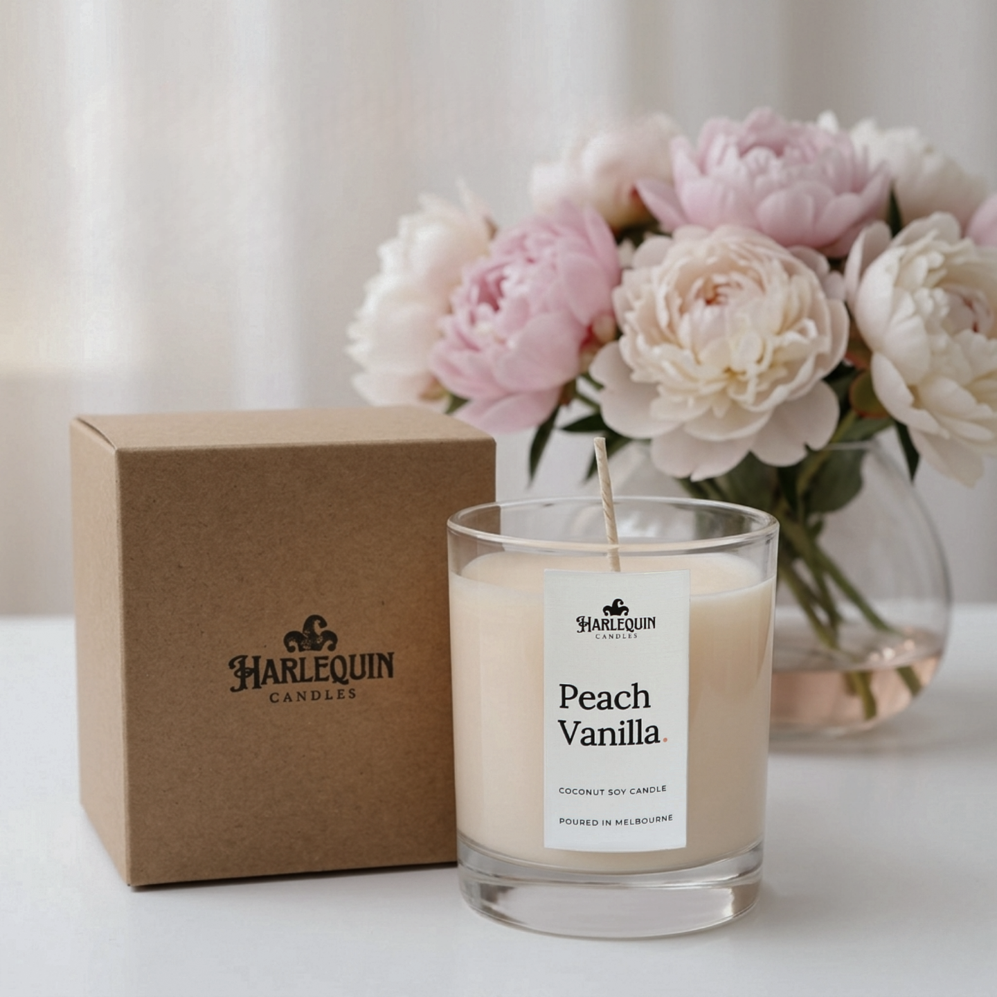 A close-up studio photograph of a white-wax Harlequin Candles "Peach Vanilla" Coconut Soy Candle in a clear glass jar, handpoured in Melbourne. The unlit candle is next to its kraft cardboard box with the brand logo, all surrounded by soft pink peonies in a vase.