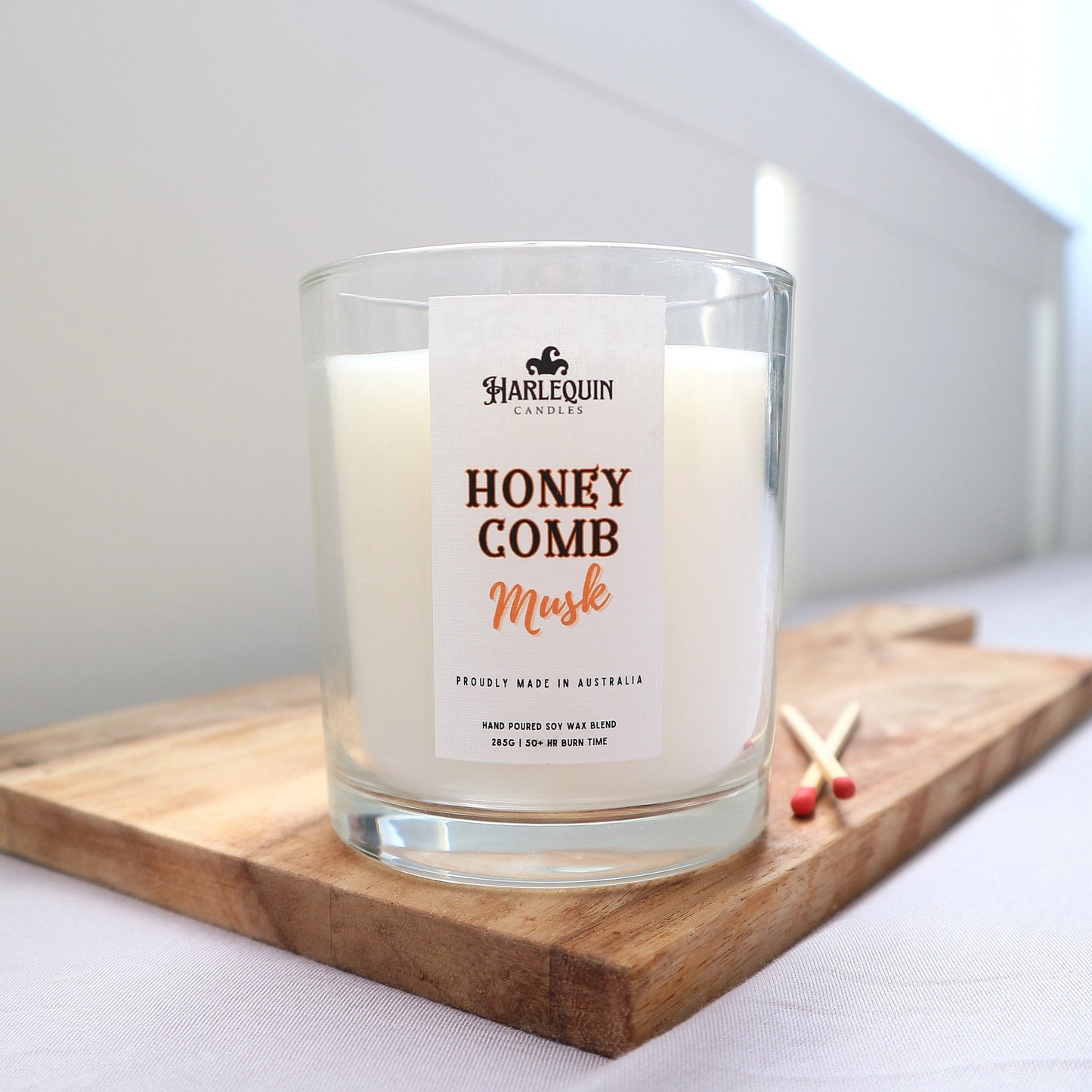 A clear glass jar candle without a lid, labeled "Honeycomb Musk" by Harlequin Candles. The candle is placed on a wooden surface.