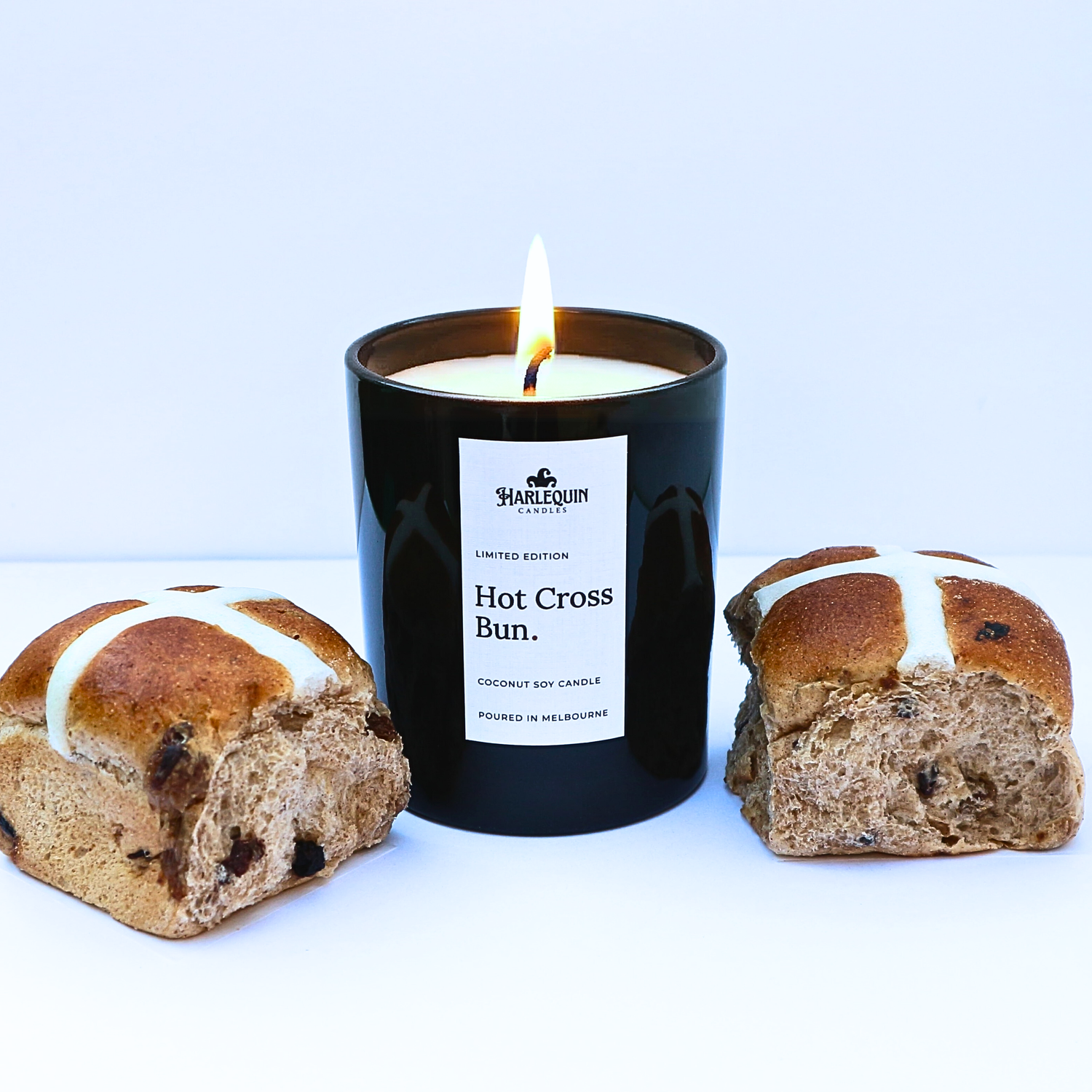 A lit Hot Cross Bun scented coconut soy candle in a sleek black jar by Harlequin Candles, styled next to fresh baked hot cross buns. Handmade and hand-poured in Melbourne, Australia.
