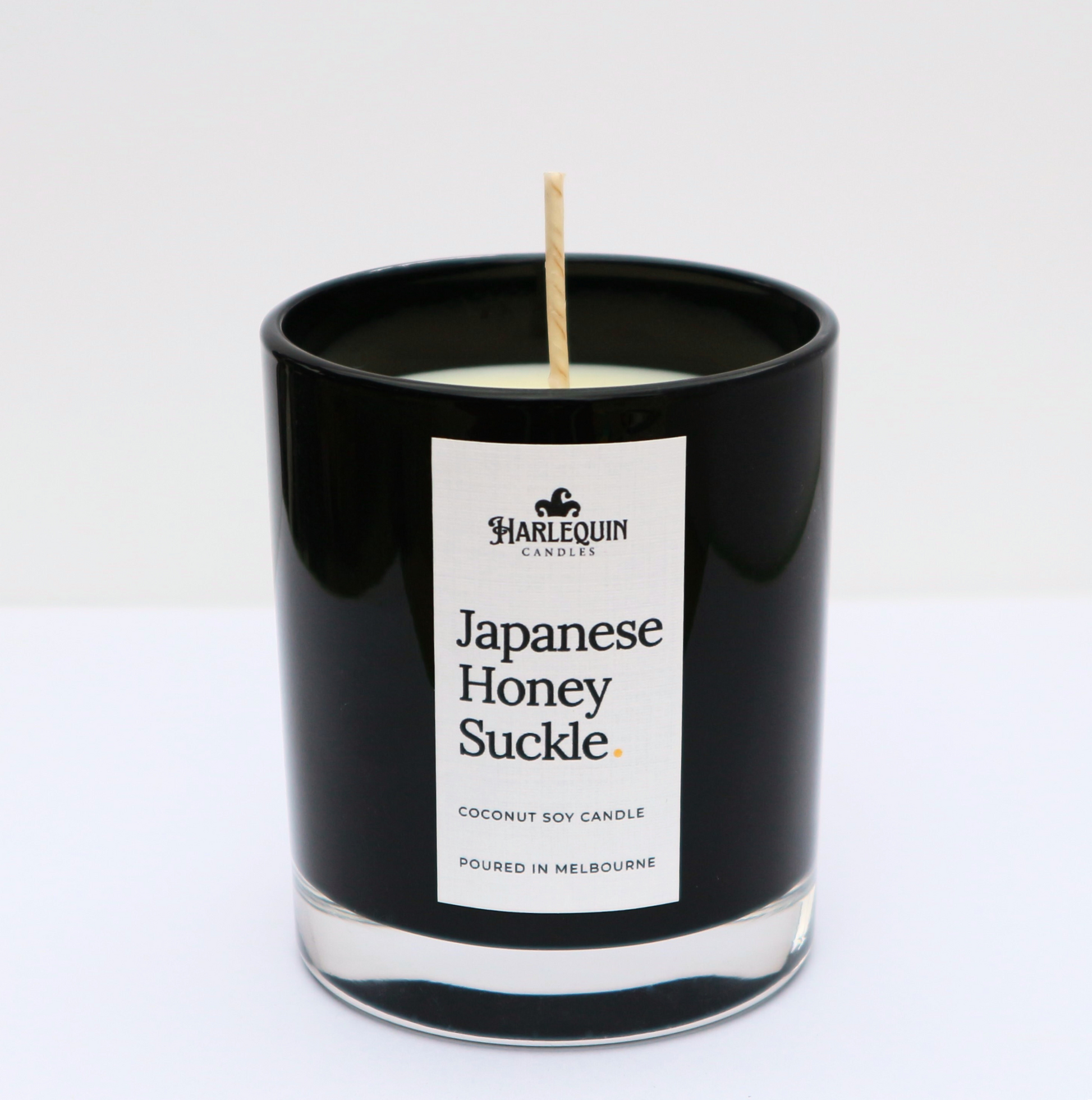 A sleek black glass jar containing a Japanese Honeysuckle coconut soy candle by Harlequin Candles. The minimalist white label features bold black text and the brand's logo, with the text "Poured in Melbourne" at the bottom against a clean, bright background.