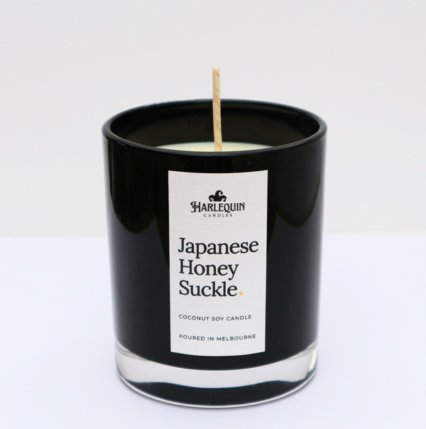 A sleek black glass jar containing a Japanese Honeysuckle coconut soy candle by Harlequin Candles. The minimalist white label features bold black text and the brand's logo, with the text "Poured in Melbourne" at the bottom against a clean, bright background.