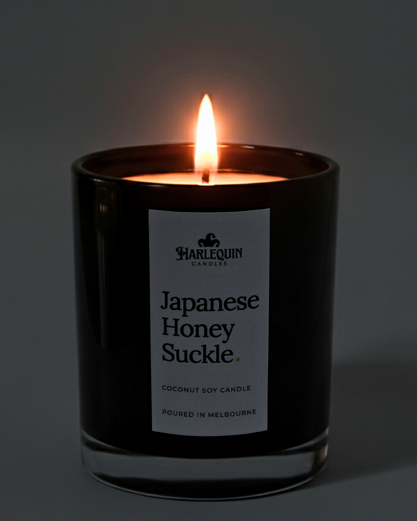 A lit Harlequin Candles premium Japanese Honeysuckle coconut soy candle in a glossy black glass jar. The warm flame illuminates the minimalist white label, which features the brand logo and the text "Poured in Melbourne" against a soft, dark background.