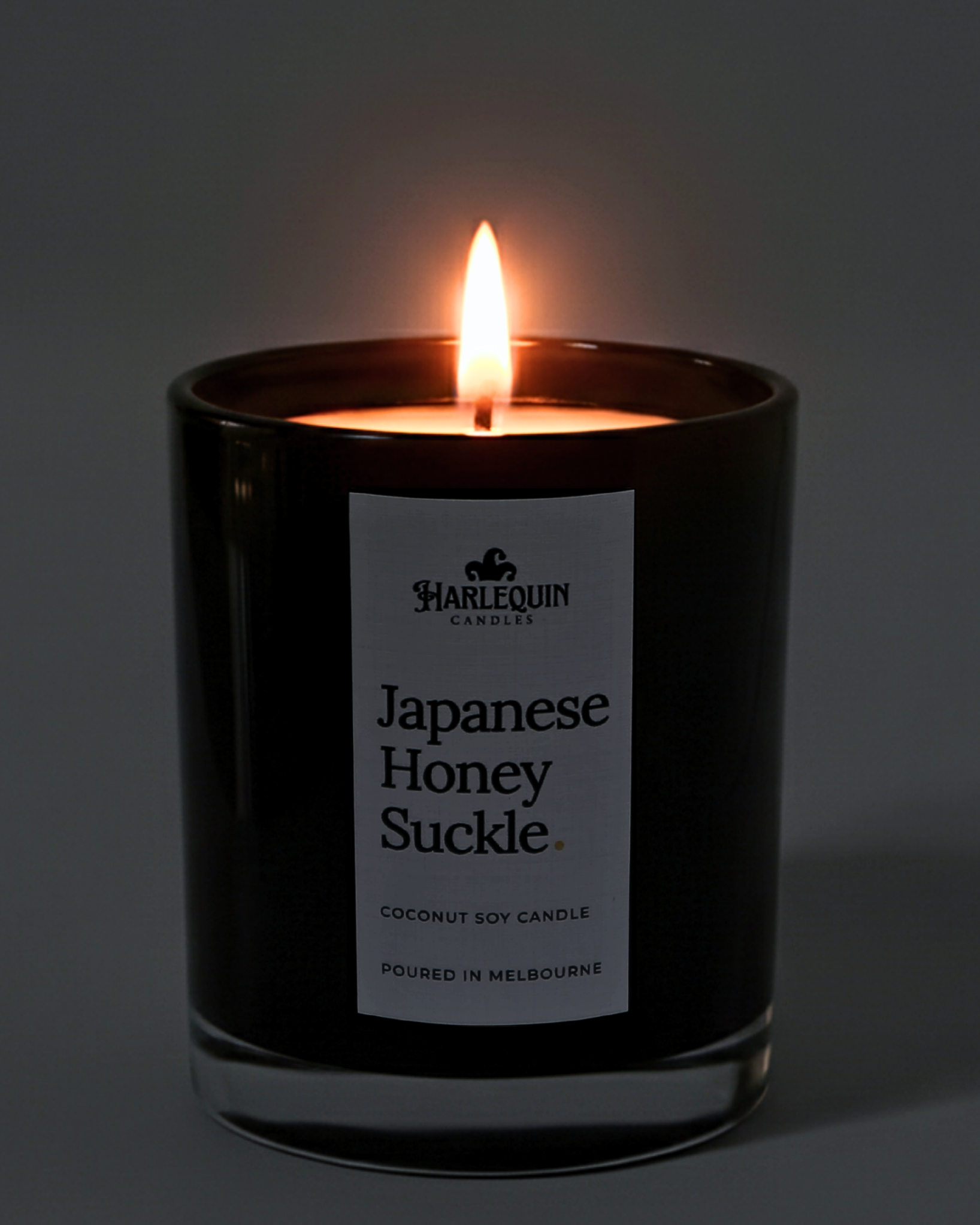 A lit Harlequin Candles premium Japanese Honeysuckle coconut soy candle in a glossy black glass jar. The warm flame illuminates the minimalist white label, which features the brand logo and the text "Poured in Melbourne" against a soft, dark background.