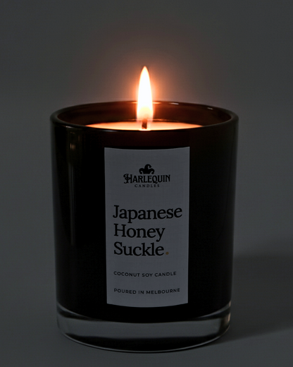 A lit Harlequin Candles premium Japanese Honeysuckle coconut soy candle in a glossy black glass jar. The warm flame illuminates the minimalist white label, which features the brand logo and the text "Poured in Melbourne" against a soft, dark background.