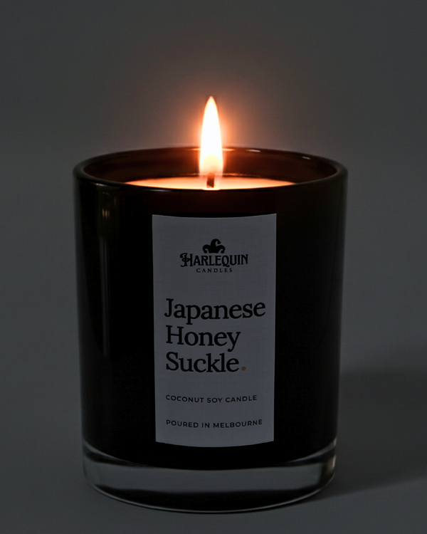 A lit Harlequin Candles premium Japanese Honeysuckle coconut soy candle in a glossy black glass jar. The warm flame illuminates the minimalist white label, which features the brand logo and the text "Poured in Melbourne" against a soft, dark background.