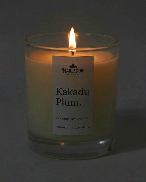 A lit Harlequin Candles luxury Kakadu Plum coconut soy candle in a clear glass jar. The warm flame illuminates the minimalist white label, which features the brand logo and the text "Poured in Melbourne" against a soft, dark background.
