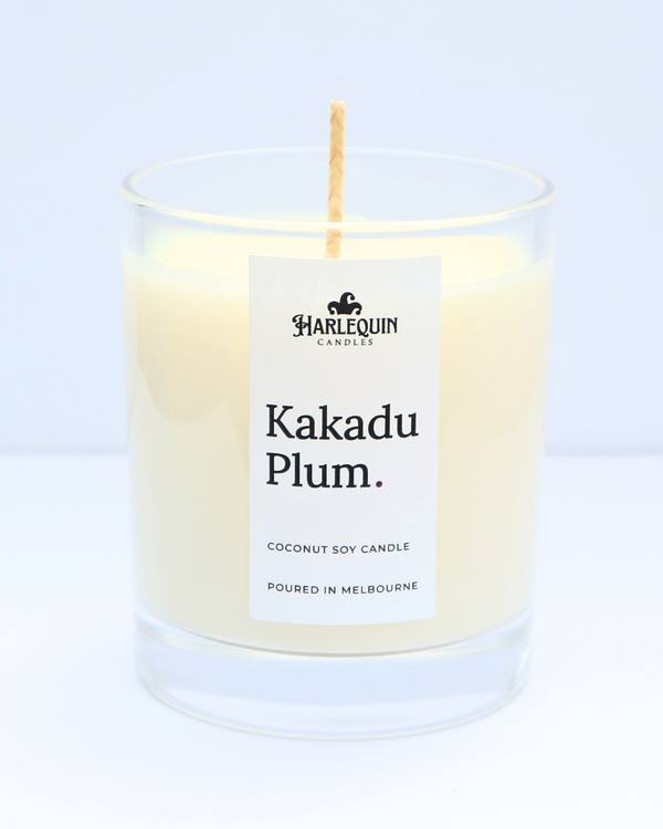 A close-up of a Kakadu Plum coconut soy candle by Harlequin Candles in a clear glass jar. The minimalist white label features bold black typography and the brand's logo, with the text "Poured in Melbourne" clearly visible at the bottom against a clean, bright background.