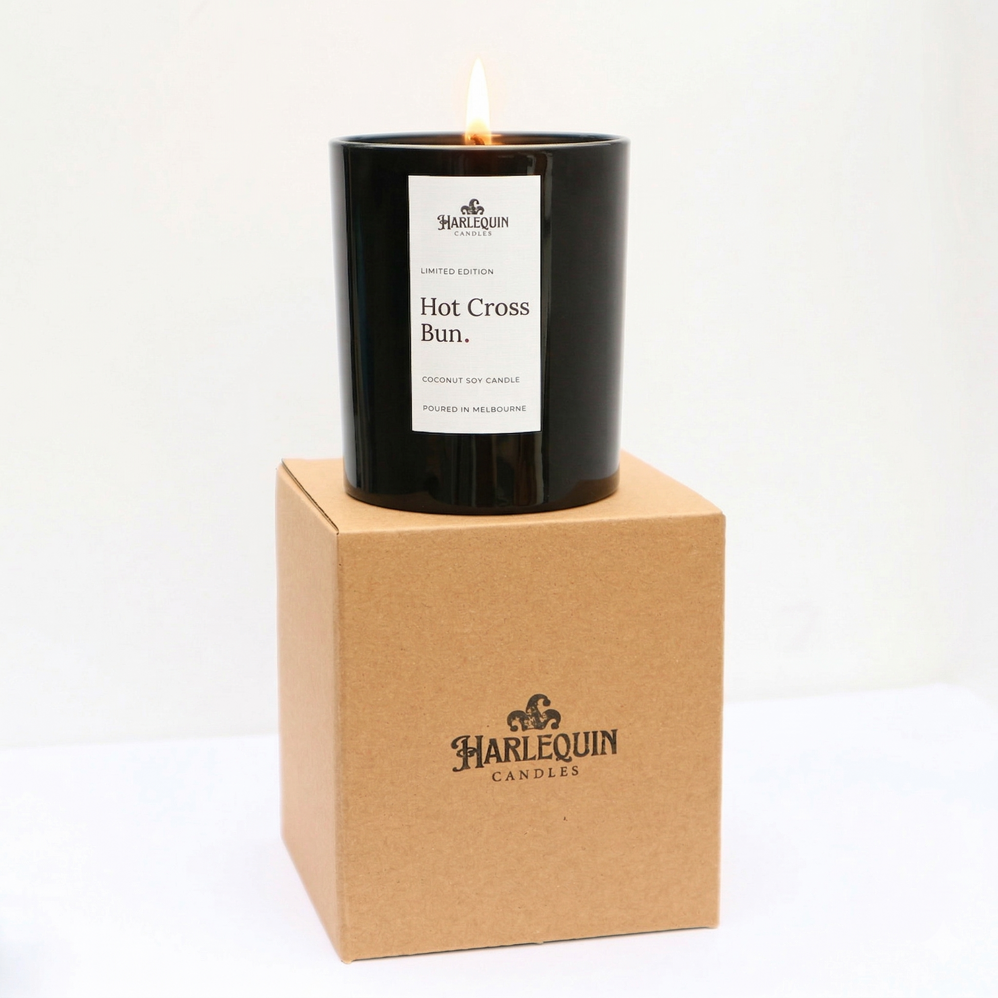 A lit Limited Edition Hot Cross Bun coconut soy candle in a glossy black jar, balanced on top of its brown kraft gift box. Both the jar and the box feature the Harlequin Candles logo, emphasizing the hand-poured Melbourne branding against a clean white background.