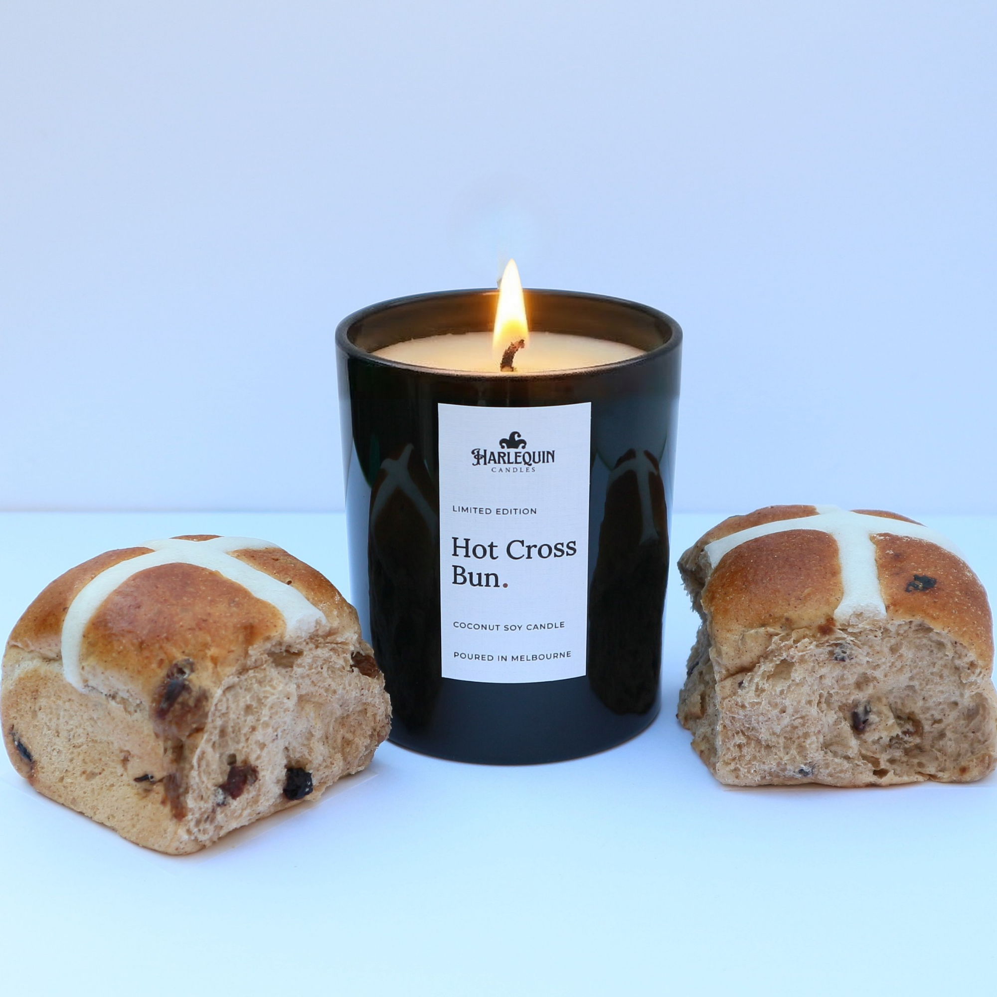 A lit Hot Cross Bun limited edition coconut soy candle by Harlequin Candles in a glossy black jar. The candle is centered between two halved, fresh hot cross buns, highlighting the fragrance's inspiration. The label clearly states "Poured in Melbourne" against a bright, clean background.
