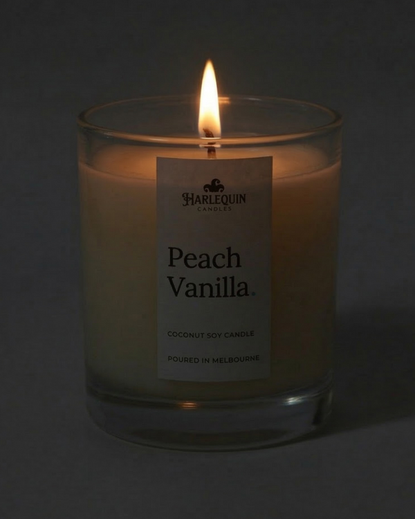A lit Harlequin Candles premium Peach Vanilla coconut soy candle in a clear glass jar. The warm flame illuminates the minimalist white label, which features the brand logo and the text "Poured in Melbourne" against a soft, dark background.
