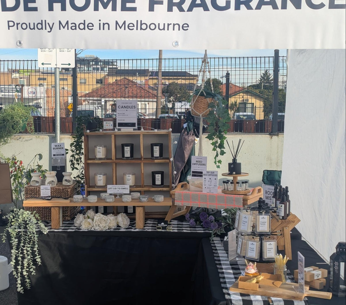 A market stall display for 'Handmade Home Fragrance' products under a white canopy banner that reads 'Proudly Made in Melbourne'. The stall features wooden shelves with small bottles, jars, and decorative items, alongside a black draped table and a checkered table with candles and reeds.