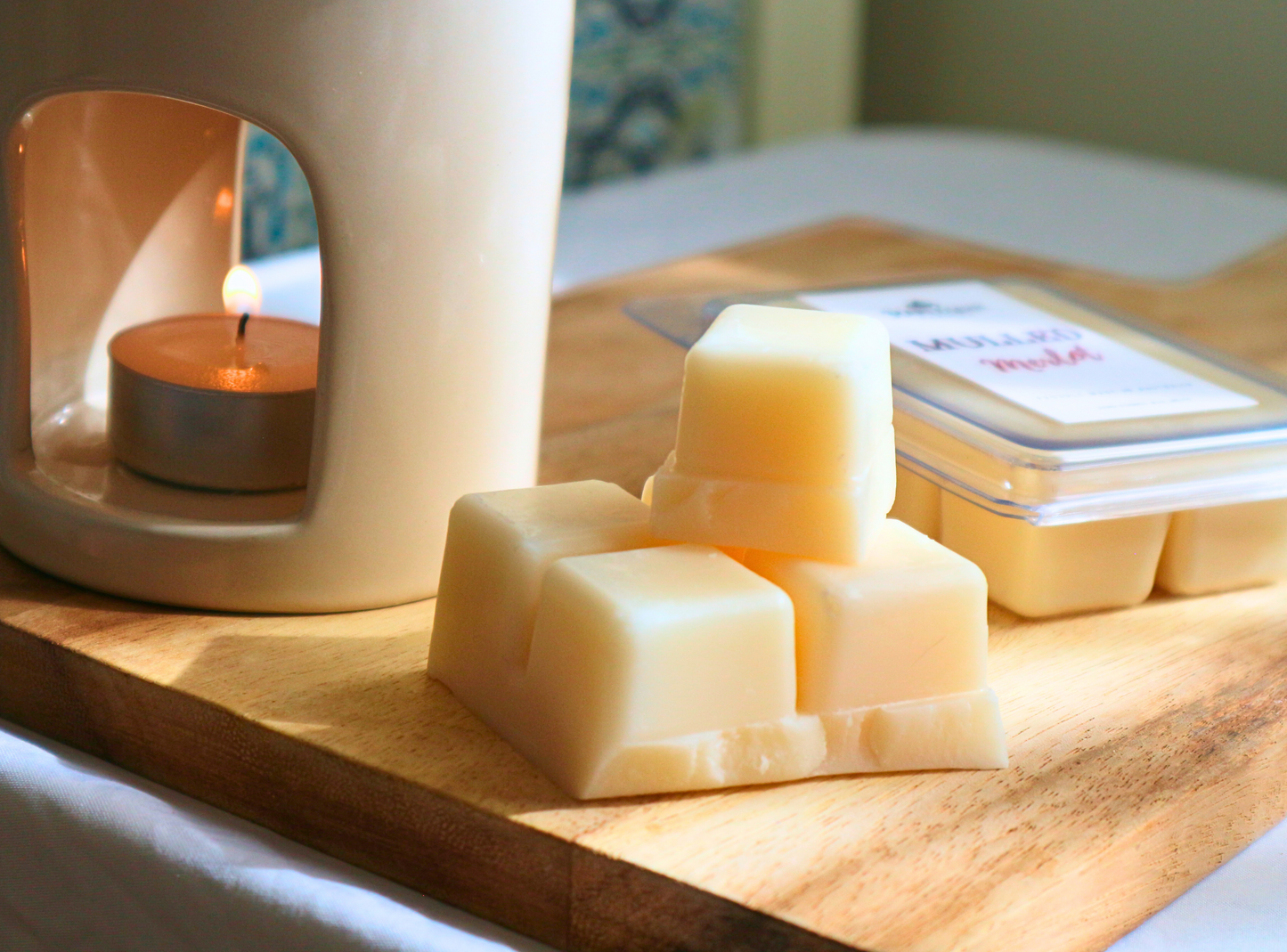 Zoom in of a stack of soy wax melt cubes next to a lit burner with a tealight and a clam-shell packaging stating Harlequin Candles. 