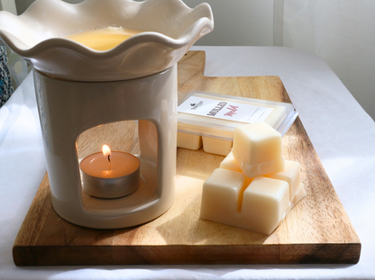 A stack of cream colored soy wax melts next to a lit burner and a clamshell package of wax melts. 